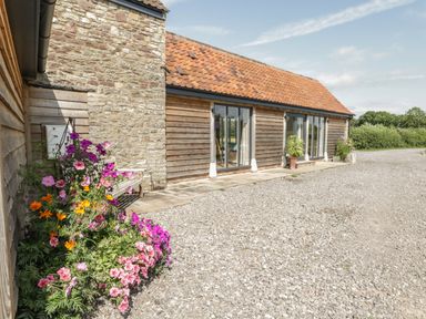 The exterior of a stone and wood barn with a tiled roof flowers near a bench and gravel driveway at Golden Valley Barn Oldbury-On-Severn