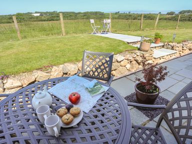 A patio table with a teapot two cups muffins an apple and a map in a garden with a stone wall and chairs at Y Bwthyn in Brynsiencyn