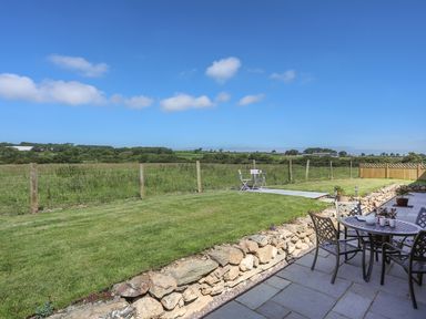 An outdoor patio area with a metal table and chairs next to a stone wall and lawn with a small seating area and fields in the background at Y Bwthyn in Brynsiencyn