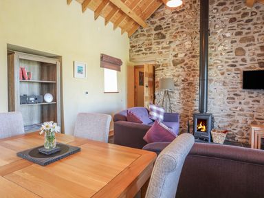 A living room with a wooden dining table chairs a sofa wood burning stove and stone wall at Y Bwthyn in Brynsiencyn