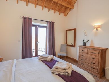 A bedroom with a bed with folded towels on a purple runner a wooden chest of drawers with a potted plant a wall-mounted light a chair a wall mirror and glass doors with curtains at Y Bwthyn in Brynsiencyn