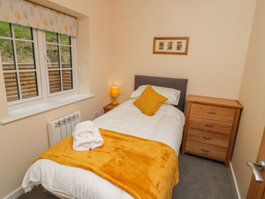 A small bedroom with a single bed a yellow blanket a pillow two folded towels a wooden chest of drawers and a window with a patterned blind at Ryedale Country Lodges - Hazel Lodge in Kirkbymoorside
