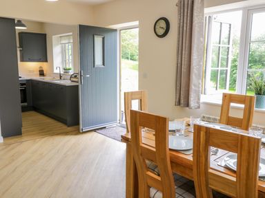 A dining area with wooden chairs and a table set with plates and glasses next to an open door and a kitchen with dark cabinets at Ryedale Country Lodges - Hazel Lodge in Kirkbymoorside