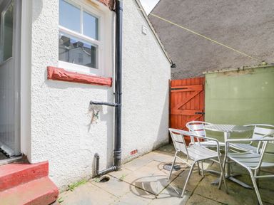 An outdoor patio area with a metal table and four chairs a white wall a black drainpipe a red wooden gate and concrete paving at Sefton House in Keswick