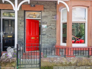 Front entrance of a building with a red door slightly open black gate and window reflecting a red car at Sefton House in Keswick