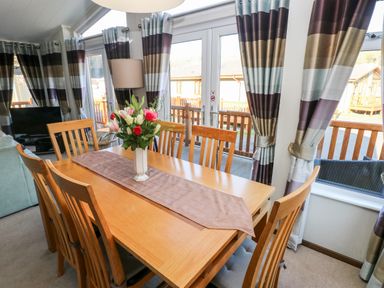 A dining area with wooden table and chairs and a flower vase near windows with striped curtains at Serendipity - Heritage Park in Stepaside near Kilgetty