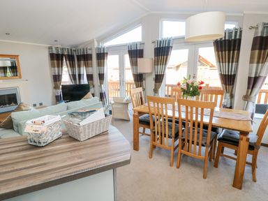 A dining area with a wooden table and six chairs next to windows with striped curtains and a living area with a sofa and television at Serendipity - Heritage Park in Stepaside near Kilgetty