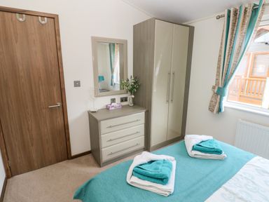 A bedroom with a wooden door dresser with mirror wardrobe bed with towels and window with curtains at Serendipity - Heritage Park Stepaside near Kilgetty