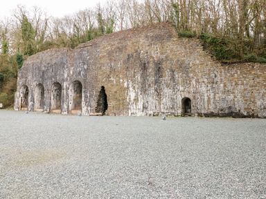 An old stone wall with multiple arched openings surrounded by gravel ground and trees in the background at Serendipity - Heritage Park Stepaside near Kilgetty