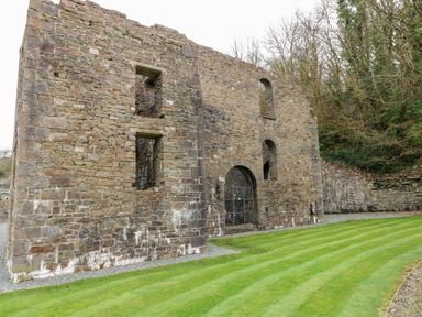 An exterior view of a stone ruin with arched windows and a gated entrance next to a green striped lawn surrounded by trees at Serendipity - Heritage Park in Stepaside near Kilgetty