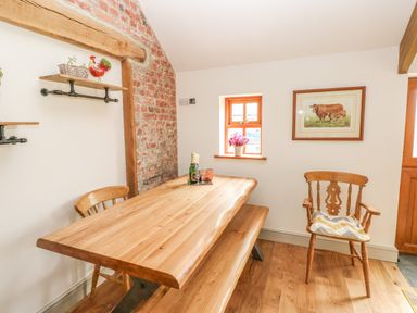 A dining area with a wooden table bench chairs a small window with flowers and a framed cow picture at The Old Farm Shop in Narberth