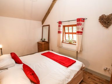 A bedroom with a bed with red cushions and blanket by a window with red curtains and a dresser with mirror at The Old Farm Shop in Narberth