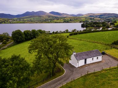 A small white cottage with a gravel driveway surrounded by green fields near a lake with mountains in the background at Lough Island Reavy Cottage Kilcoo Mourne Mountains Newry County Down