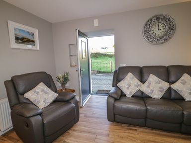 A living room with two grey leather sofas with floral cushions an open door showing outside a grassy area a clock on the wall and a framed picture at Lough Island Reavy Cottage in Kilcoo Mourne Mountains Newry County Down
