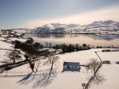 A snow-covered landscape with a small house near a lake and mountains in the background at Lough Island Reavy Cottage in Kilcoo Mourne Mountains Newry County Down