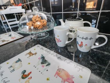 A kitchen counter with a glass cake stand holding cookies two mugs and a teapot with farm animal illustrations at The Old Well Bungalow - Ty Mawr Farm in Cardiff