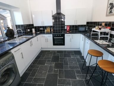 A kitchen with white cabinets black tiled backsplash a sink oven microwave and two wooden stools at The Old Well Bungalow - Ty Mawr Farm Cardiff