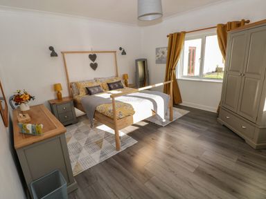 A bedroom with a wooden bed, bedside tables, a dresser with a mirror, a wardrobe, a full-length mirror, and a window with yellow curtains at The Old Well Bungalow - Ty Mawr Farm in Cardiff