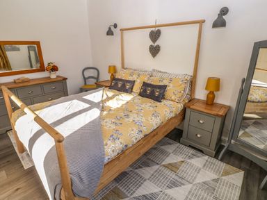 A bedroom with a wooden bed frame yellow floral bedding bedside tables with lamps a dresser with a mirror a gray chair and a floor mirror at The Old Well Bungalow - Ty Mawr Farm in Cardiff