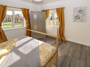A bedroom with a wooden bed with yellow floral bedding a grey wardrobe with drawers two windows with mustard curtains and a wall painting at The Old Well Bungalow - Ty Mawr Farm in Cardiff
