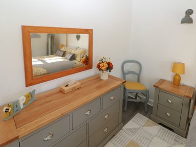 A bedroom corner with a wooden dresser topped with flowers and a tissue box a chair with a yellow seat and a small wooden nightstand with a yellow lamp at The Old Well Bungalow - Ty Mawr Farm Cardiff