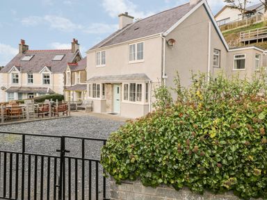 A two-story house with a gravel driveway and a black gate next to a bush and neighboring houses at Castell in Abersoch