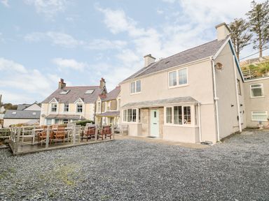 A detached two-story house with a front porch and a gravel driveway with wooden outdoor seating on a deck next to the house at Castell in Abersoch