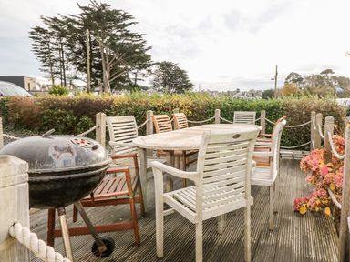An outdoor patio with a wooden table surrounded by chairs and a charcoal barbecue grill on a deck with plants and trees at Castell in Abersoch