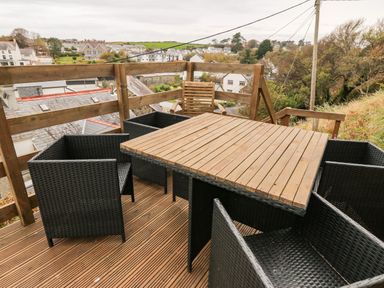 An outdoor wooden deck with a wooden table and four black wicker chairs overlooking houses and green hills at Castell in Abersoch