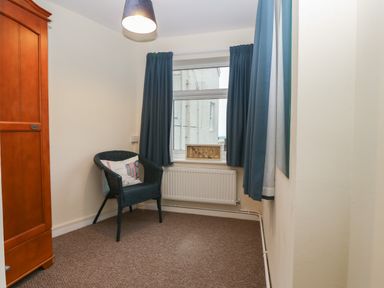 A small room with a black chair with cushion a wooden wardrobe blue curtains and a window at Castell in Abersoch