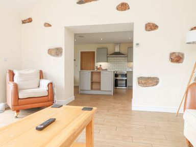 A living room with a leather armchair and wooden table looking towards a kitchen with cabinets and an oven at The Cottage in Llaniestyn
