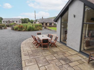An outdoor patio area with a table and eight wooden chairs next to a modern house with large windows at The Cottage in Llaniestyn