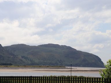 A coastal landscape with hills in the background a body of water and a sandy shore with a black picket fence and a lamp post in the foreground at Sunset Sands in Deganwy