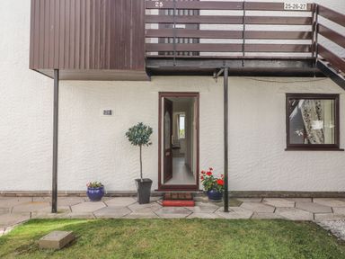 A ground floor entrance door with a doormat between two flower pots and a small tree in a pot outside a residential building at Sunset Sands in Deganwy