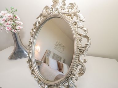 An ornate oval mirror reflecting a bed with pillows and a bedside lamp with a flower vase on a table