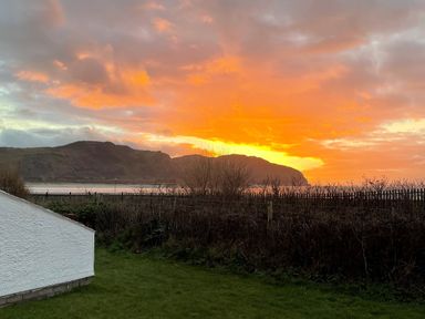 A sunset with orange sky behind mountains and a grassy yard with a white wall at Sunset Sands in Deganwy
