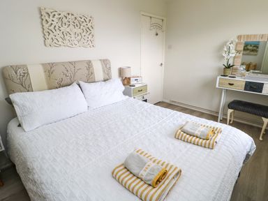 A bedroom with a double bed white bedding and folded towels on the bed a bedside table with a lamp a dressing table with a mirror and a stool at Sunset Sands in Deganwy