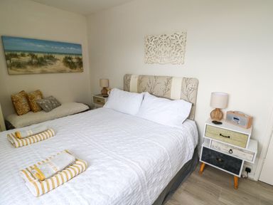 A bedroom with a bed with white bedding two bedside tables lamps and a painting of a beach at Sunset Sands in Deganwy