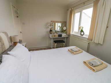 A bedroom with a bed with white bedding a dressing table with a mirror and a window with curtains at Sunset Sands in Deganwy