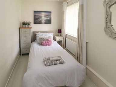 A small bedroom with a single bed bedside table lamp tall chest of drawers and a window with curtains at Sunset Sands in Deganwy