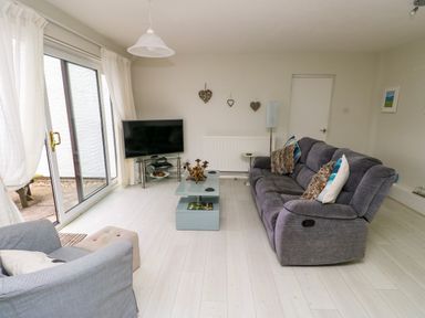 A living room with a gray sofa a blue armchair a glass coffee table a TV on a stand and sliding glass doors at Sunset Sands in Deganwy