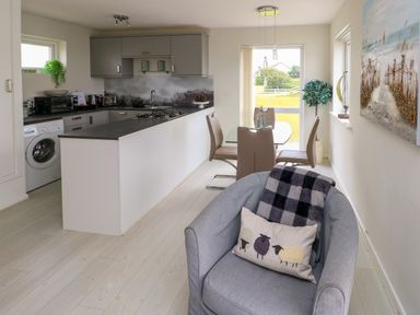 A kitchen and dining area with a gray armchair in the foreground and glass dining table with four chairs at Sunset Sands in Deganwy