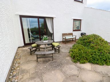 An outdoor patio with a wooden picnic table with green cushions a bench a bush and sliding glass doors at Sunset Sands in Deganwy