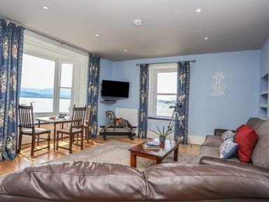 A living room with blue walls floral curtains wooden chairs a rocking horse a telescope and a brown leather sofa at Gadlys House - Waters Edge in Beaumaris