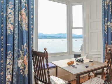 A corner dining area with a wooden table and chairs by a bay window overlooking a body of water with boats and mountains at Gadlys House - Waters Edge Beaumaris