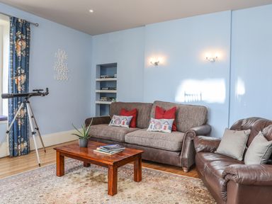 A living room with blue walls a brown sofa and armchair wooden coffee table telescope and patterned curtains at Gadlys House - Waters Edge Beaumaris