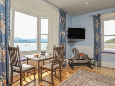 A dining area with a small table and two chairs by bay windows showing a water view with boats and mountains at Gadlys House - Waters Edge in Beaumaris