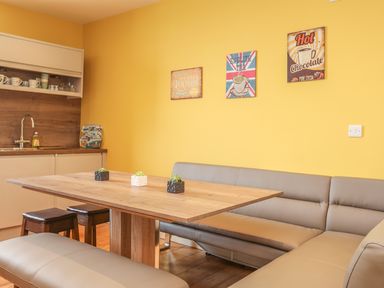 A kitchen dining area with a wooden table gray seating stools and yellow walls with decorative signs at Gadlys House - Waters Edge in Beaumaris