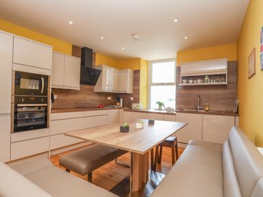 A kitchen with wooden table and bench seating with white cabinets and yellow walls at Gadlys House - Waters Edge in Beaumaris