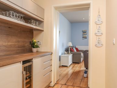 A kitchen area with glassware and a wooden countertop leading to a living room with a sofa and side table at Gadlys House - Waters Edge in Beaumaris